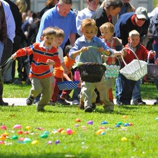 A group of young children holding Easter baskets run across a lawn covered with plastic eggs while their parents cheer behind them.