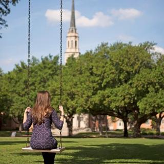 Back of female TCU student sitting in a metal swing with a cluster of trees and the TCU chapel in the background.