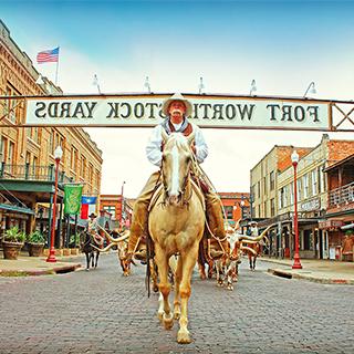 A cowboy in customary western attire rides a brown and white horse underneath a "Fort Worth Stock Yards" sign that spans across a street lined with old western buildings
