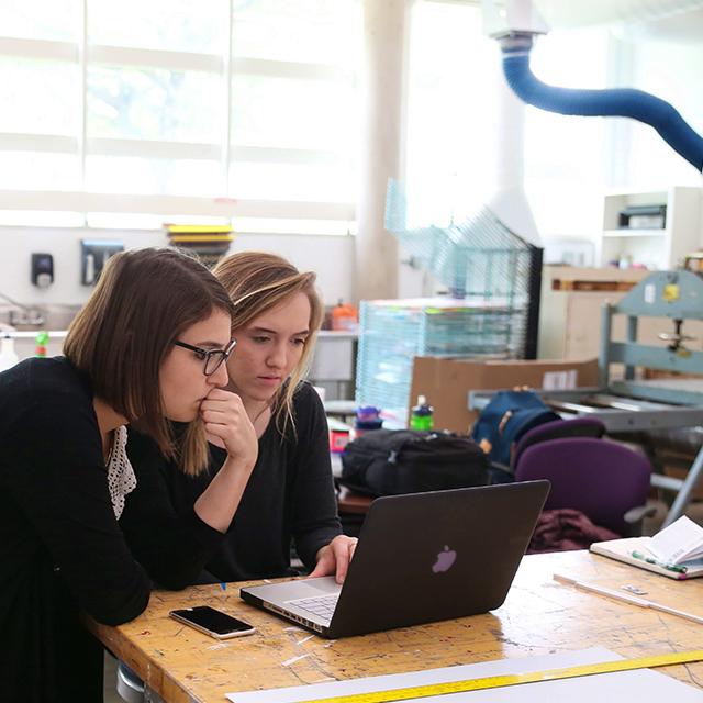 Two female TCU students view a laptop in the printmaking studio