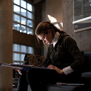 A young women sits alone and looks down intently at her book in the sunlit lobby of TCU's Moudy Hall North.